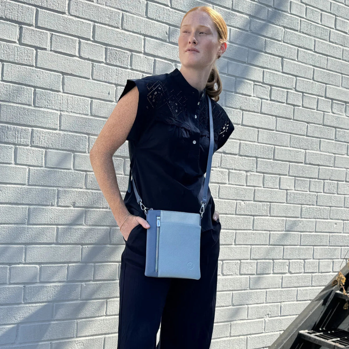 Woman in classic black clothes and blue crossbody bag, standing by a white brick wall.