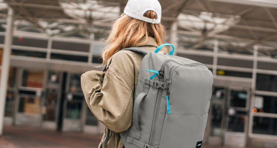 Woman walking towards doors of a train station carrying a Pacsafe® GO 44L Anti-theft Carry-on Backpack in stone grey.