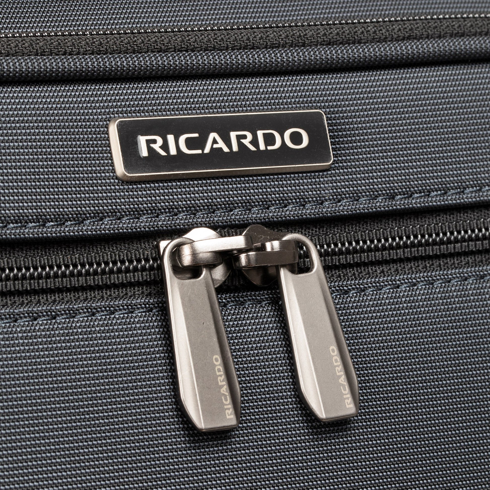Close-up of Ricardo Beverly Hills Montecito 3.0 Large Check-In Spinner zipper and logo on dark grey fabric.