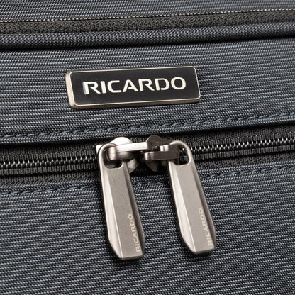 Close-up of Ricardo Beverly Hills Montecito 3.0 Large Check-In Spinner zipper and logo on dark grey fabric.