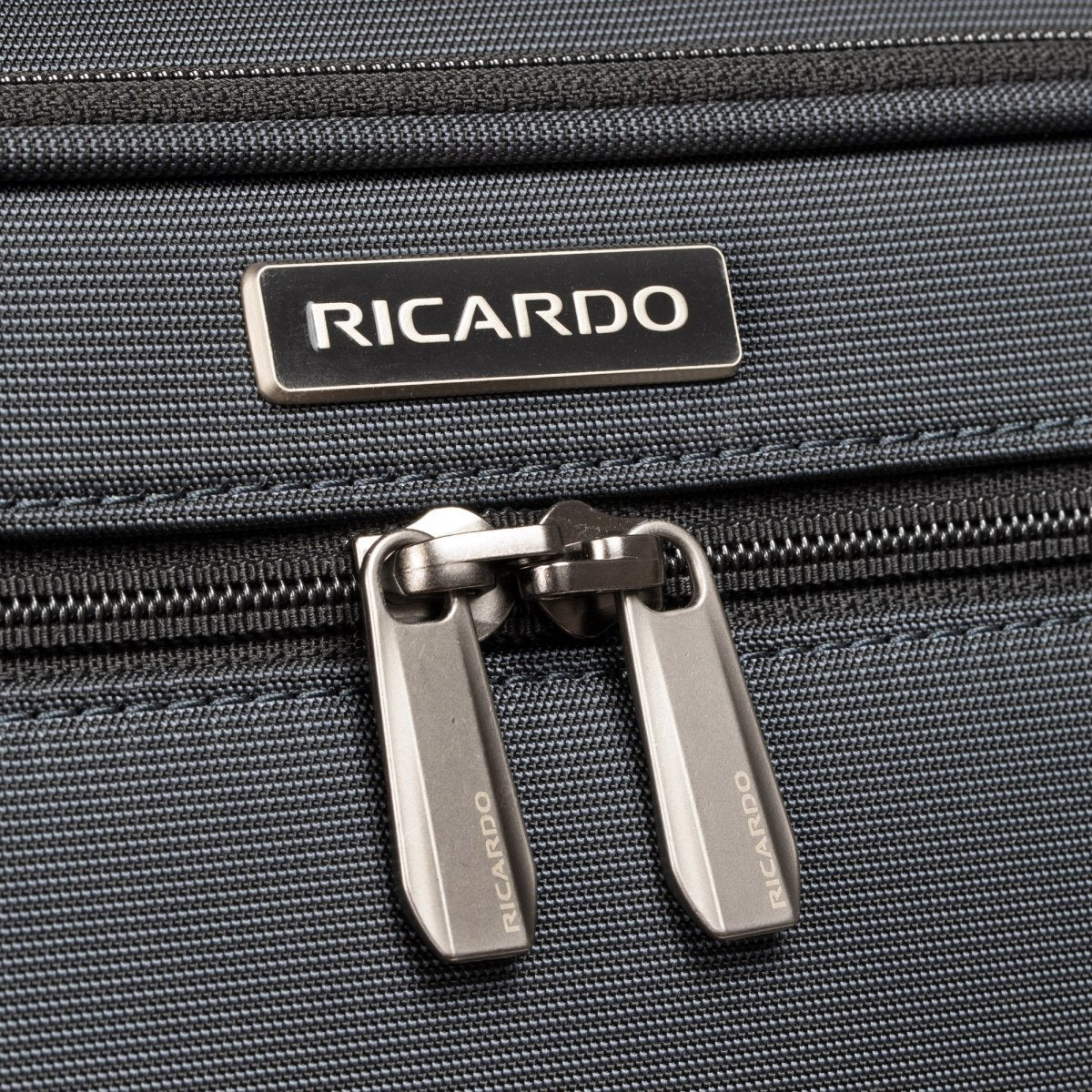 Close-up of Ricardo Beverly Hills Montecito 3.0 Large Check-In Spinner zipper and logo on dark grey fabric.