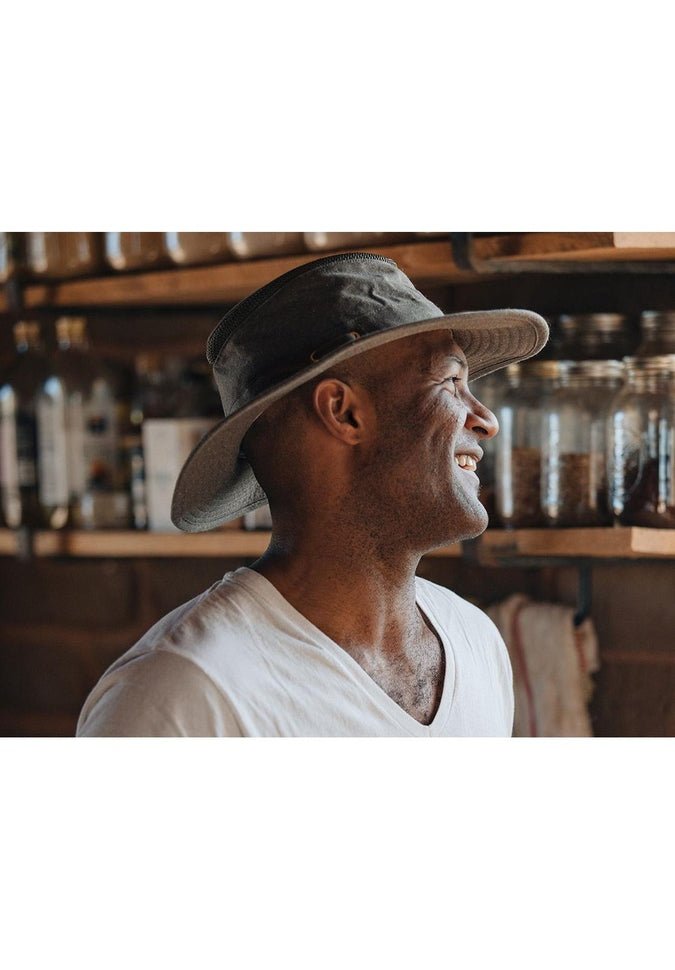 A man wearing the Tilley TMH55 Mash-Up Airflo® wide-brimmed hat, made from UPF 50+ sun protective fabric, smiles as he stands in a room with shelves filled with jars and bottles.