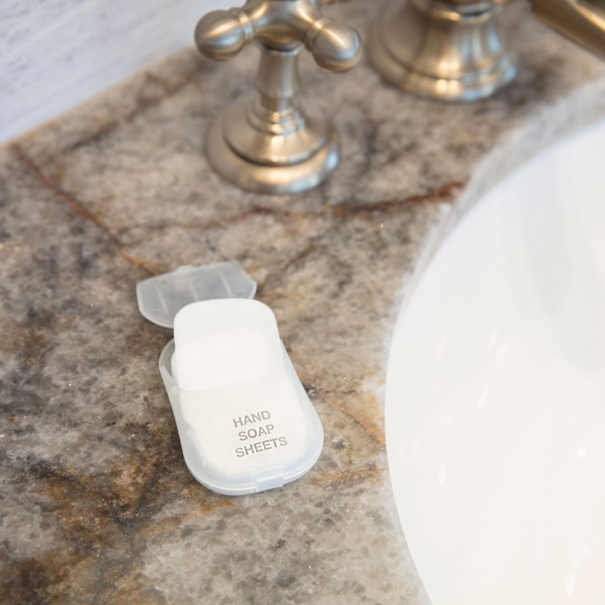 A compact plastic container labeled "Travelon Hand Soap Toiletry Sheets" on a marble countertop beside a sink faucet.