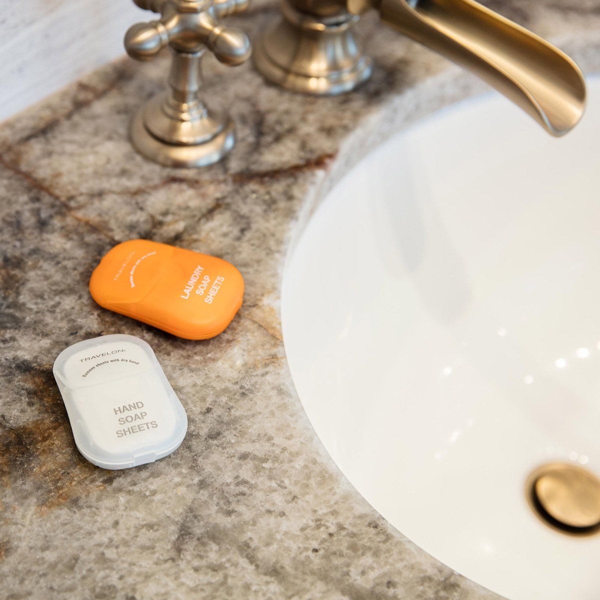 Two containers of soap sheets, one labeled "Hand Soap Sheets" and the other "Travelon Laundry Soap Toiletry Sheets" by Travelon, placed on a marble countertop beside a sink with a brass faucet.