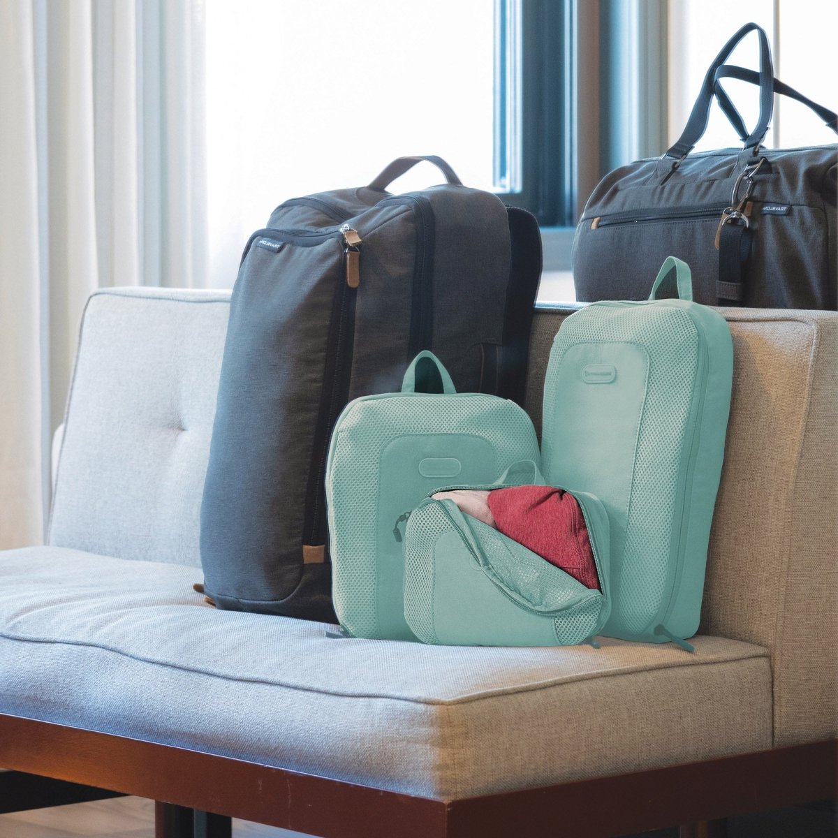 The Travelon Set of 3 Packing Cubes and other stylish travel bags and accessories are neatly arranged on a gray cushioned bench and a dark wood table.