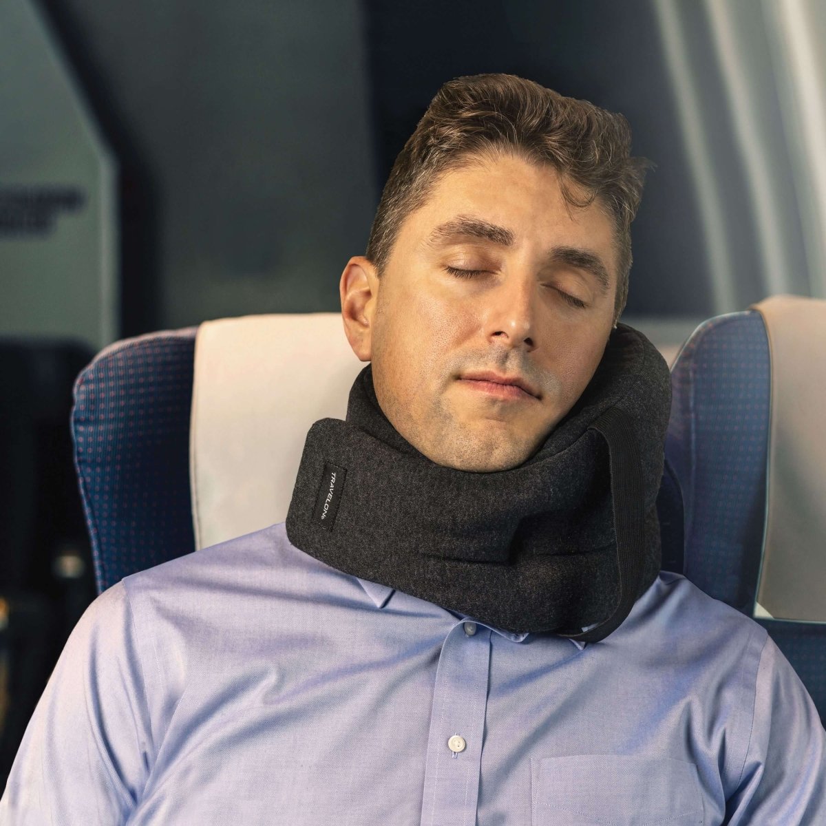 A person peacefully sleeping on a plane, nestled in an aisle seat, using a Travelon SOMNiWRAP Travel Pillow in black, designed ergonomically for optimal neck support.