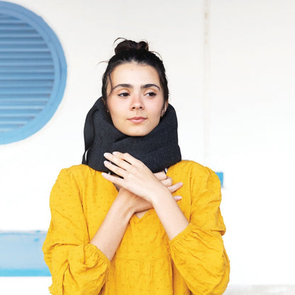 A person with dark hair styled in a bun is wearing a black Travelon SOMNiWRAP Travel Pillow, known for its ergonomic neck support, along with a yellow long-sleeve top. They stand in front of a blue circular vent on a white wall.