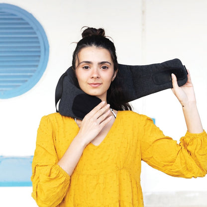 A person in a yellow dress holds a black Travelon SOMNiWRAP Travel Pillow around their shoulders in front of a blue shutter.