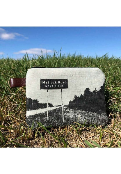 A small interlined coin purse from Winnipeg North of Fargo, featuring a black and white road sign design, is standing upright on grass under a blue sky.